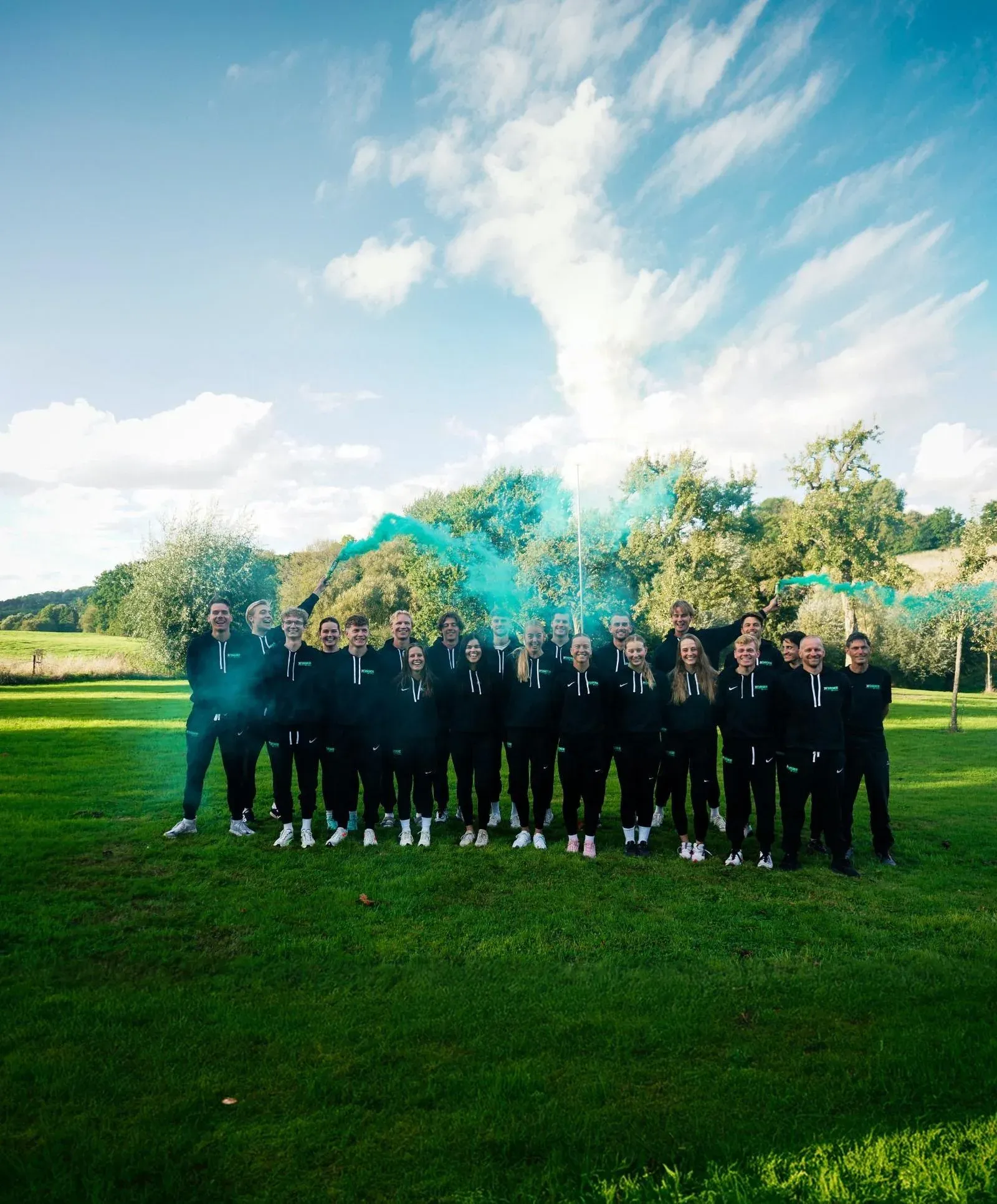 De ploeg van Schaatsteam Next | Utibo in Next-hoodies op een grasveld, met groene rook